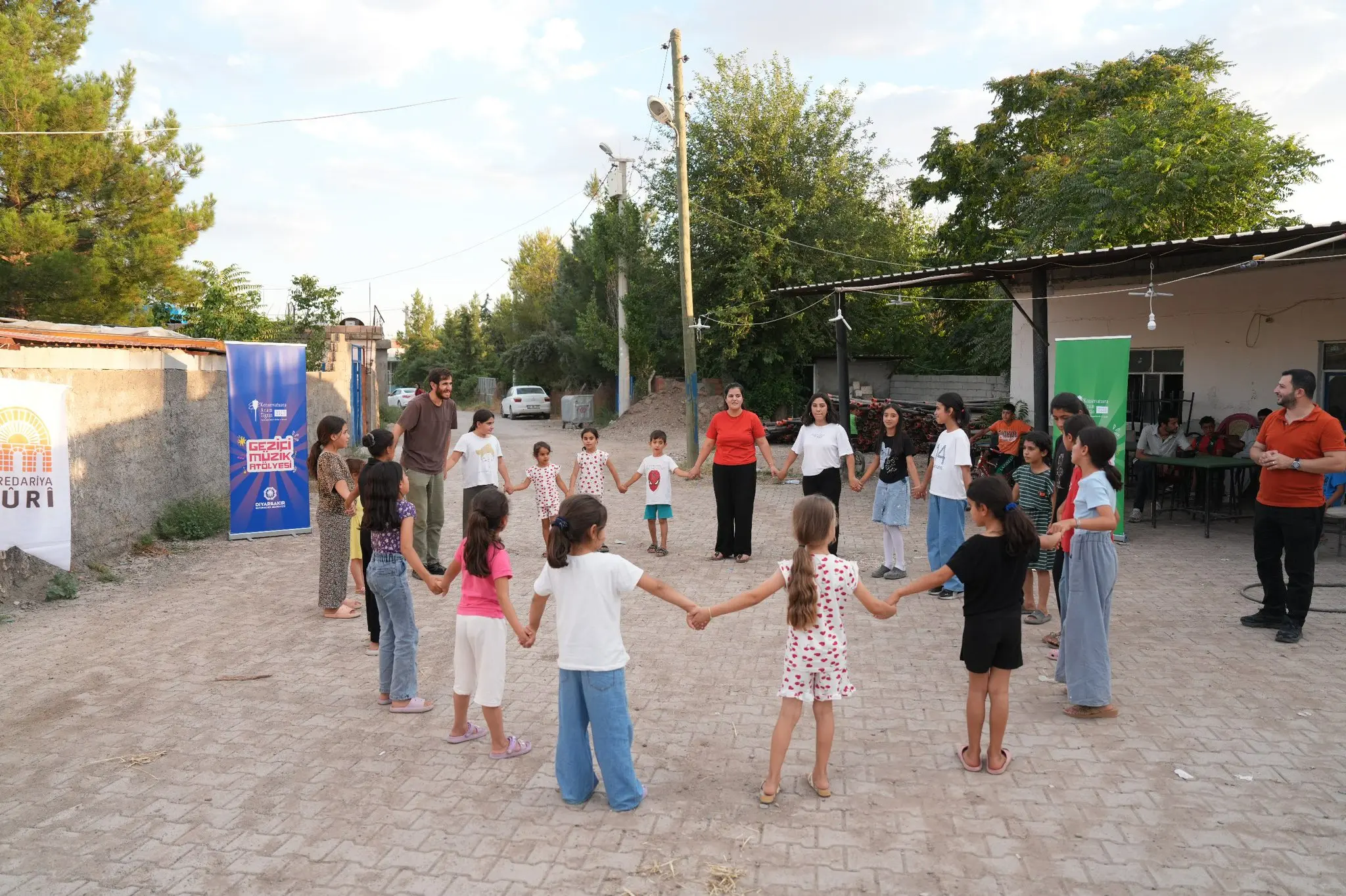 Fotoğrafta, Sur ilçesine bağlı Kervanpınar Mahallesi’nde gerçekleştirilen Gezici Müzik Atölyesi etkinliğinde çocuklar ve eğitmenler geniş bir halka oluşturarak el ele tutuşmuş şekilde oyun ve ritim çalışması yapıyor. Etkinlik açık havada, köy meydanında düzenlenmiş. Çocukların neşeli ve etkinliğe aktif katılım gösterdiği görülürken, eğitmenler de oyun ve müzik etkinliğini yönlendiriyor.
Sol tarafta Sur Belediyesi ile Diyarbakır Büyükşehir Belediyesi Aram Tigran Kent Konservatuvarına ait “Gezici Müzik Atölyesi” afişleri yer alıyor. Arka planda köy evleri, ağaçlar ve etkinliği izleyen mahalle sakinleri görülüyor. Doğal ve samimi atmosferi yansıtan kare, çocukların müzik aracılığıyla bir araya geldiği, dayanışma ve birlikte üretim duygusunun öne çıktığı anı yansıtıyor.