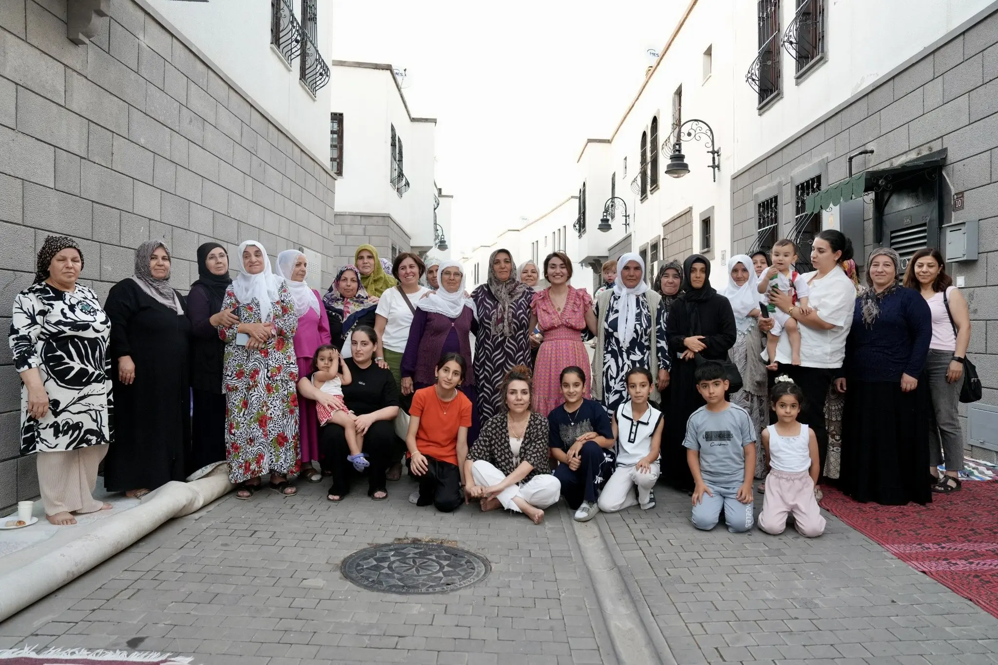 Fotoğrafta, dar ve taş döşeli bir sokakta gerçekleştirilen toplu bir buluşma görülüyor. Beyaz ve bazalt taş mimarisiyle çevrili sokakta farklı yaş gruplarından çok sayıda kadın ve çocuk bir araya gelerek objektife poz veriyor.
Grubun orta bölümünde ayakta duran kadınlar yan yana sıralanırken, ön tarafta diz çöken veya yere oturan çocuklar ve birkaç yetişkin bulunuyor. Katılımcılar kameraya gülümseyerek bakıyor ve samimi bir atmosfer yansıtıyor. Bazı kadınlar geleneksel desenli elbiseler ve başörtüleri kullanırken, bazıları günlük kıyafetler tercih etmiş. Çocuklar ise renkli ve rahat kıyafetleriyle ön sırada yer alıyor.
Fotoğrafın sağ tarafında bir kadının kucağında küçük bir çocuk bulunurken, giriş önüne serilmiş kırmızı desenli halı dikkat çekiyor. Sol tarafta ise duvar kenarında rulo hâlinde bir halı ve yerde çay bardağı görülüyor. Arka plandaki tarihi dokuya sahip binalar, ferforje balkonlar ve sokak lambaları fotoğrafa estetik bir görünüm kazandırıyor.