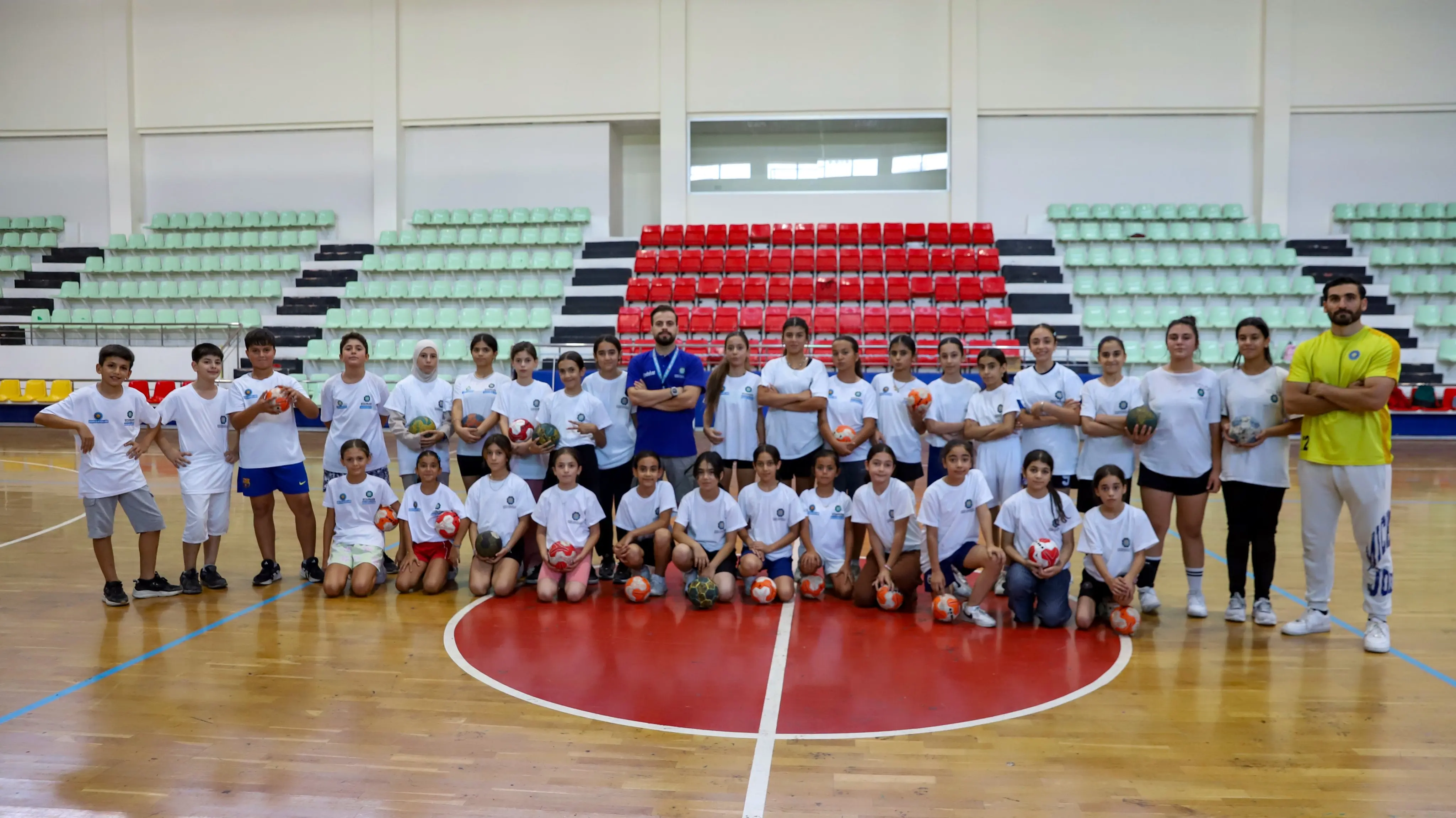 Spor salonunda, basketbol sahasının ortasında yaklaşık 30 çocuk ve 3 yetişkin birlikte duruyor. Çocukların bir bölümü ayakta, bir bölümü önde diz çökerek poz vermiş; çoğunun elinde hentbol topu bulunuyor. Çocuklar beyaz tişört ve şort giyiyor. Arkada yeşil ve kırmızı tribün koltukları yer alıyor.