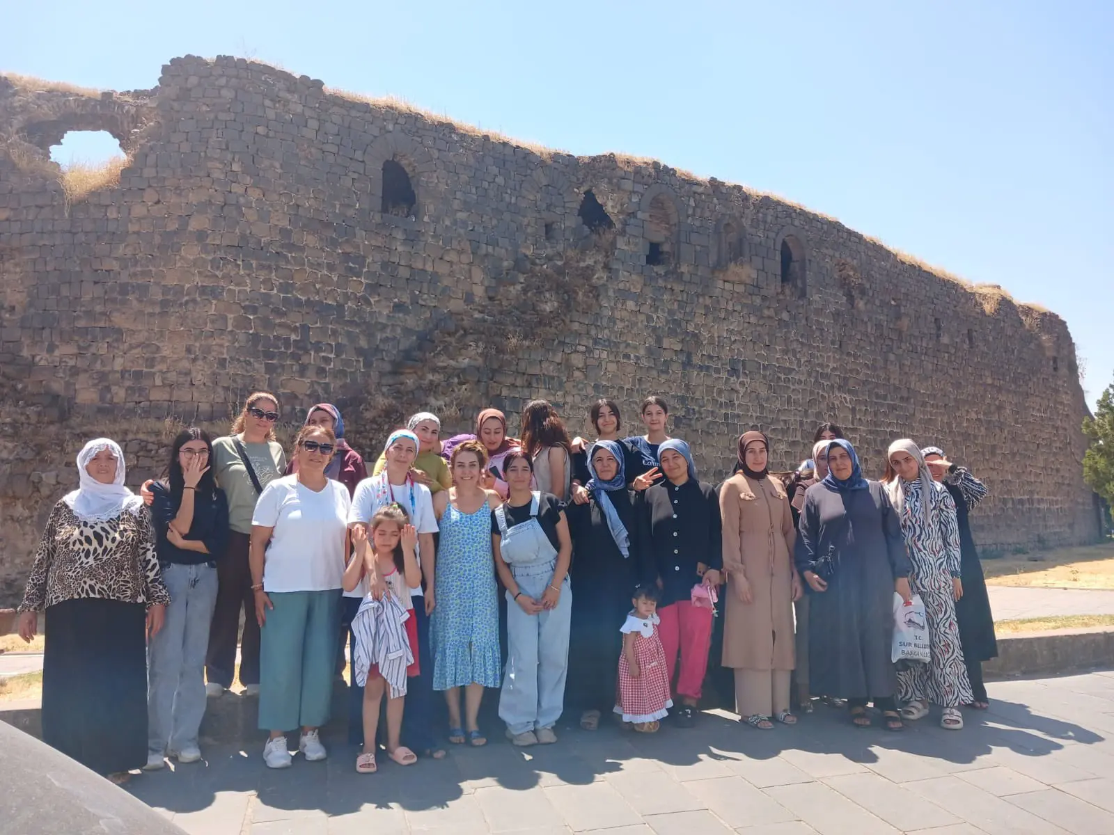 Fotoğrafta açık havada, geniş bir yüzme havuzunda gerçekleştirilen bir yüzme eğitimi görülüyor. Havuzda çoğunluğu çocuklardan oluşan bir grup, pembe ve mavi yüzme boneleriyle birlikte eğitmenlerinin eşliğinde havuz kenarında sıralanmış durumda. Bazı çocuklar havuzun kenarına tutunarak çalışırken, eğitmenler de onları yönlendiriyor.
Havuz çevresinde şezlonglar, taş duvarlar ve yeşil alanlar bulunuyor. Arka planda ise müstakil evler ve ağaçlar görülüyor. Güneşli ve açık bir havada gerçekleşen eğitim, çocukların yüzme öğrenirken keyifli ve hareketli vakit geçirdiği sıcak bir ortamı yansıtıyor.